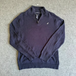 Mörkblå stickad tröja från Nautica - Mörkblå stickad tröja från Nautica med halv dragkedja och liten broderad logga på bröstet. Tillverkad i 100% bomull och har ribbade muddar vid ärmslut och nederkant. Perfekt för lager-på-lager och en stilren look.