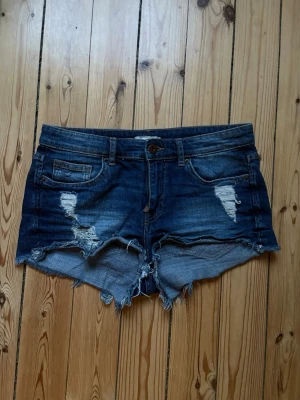 Mörkblå jeansshorts - Snygga mörkblå jeansshorts med slitna detaljer och fransig kant. Klassisk femficksmodell med knapp och dragkedja framtill. Perfekta för en avslappnad stil under varma dagar.