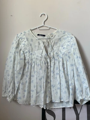 Blommig vit blus från Gina Tricot - Romantisk vit blus med ljusblått blommönster från Gina Tricot. Blusen har rund halsringning med knytband, dekorativa veck framtill och knappar. Långärmad modell i luftig bomull, perfekt för en söt och avslappnad stil.