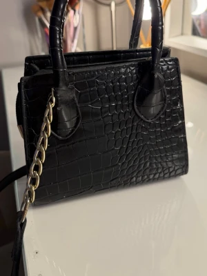 Svart croco handväska med kedja - Stilren svart handväska i croco-mönstrat konstläder med två handtag och en axelrem med guldfärgad kedjedetalj. Väskan har en fyrkantig form och dragkedja upptill. Perfekt accessoar för att lyfta din outfit.