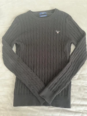 Svart kabelstickad tröja Gant - Kabelstickad svart tröja från Gant med rund halsringning och broderad logotyp på bröstet. Tillverkad i stretchig bomull för extra komfort. Långa ärmar och ribbade muddar vid ärmslut och nederkant.