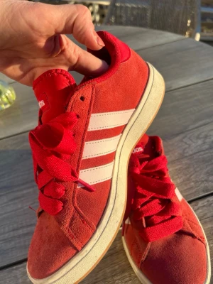 Röda Adidas sneakers med vita ränder - Snygga röda Adidas sneakers i mocka med klassiska vita ränder på sidorna och vit sula. Skorna har röda skosnören och Adidas-logga bak på hälen. Perfekta för dig som vill sticka ut med färg och sportig stil. Använda ett fåtal ggr. För små. Innermått 25 cm.