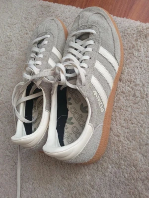 Adidas Spezial grå sneakers - Snygga Adidas Spezial sneakers i grå mocka med vita detaljer och klassiska tre ränder på sidorna. Skorna har vit hälkappa, vita skosnören och en gummisula i naturfärg. Perfekta för en stilren och sportig look.