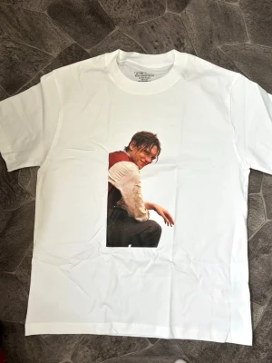 Vit Jamie campbell bower t-shirt med tryck - Säljer en vit t-shirt med ett stort tryck framtill som föreställer en person i sittande pose. T-shirten har klassisk passform och rund hals. Perfekt för dig som gillar streetwear och vill sticka ut med en unik print.