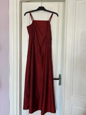 Vinröd lång klänning med smala axelband - Snygg vinröd långklänning med smala axelband och lätt glansigt tyg. Klänningen har en rak passform och draperad detalj framtill. Perfekt för dig som gillar minimalistisk och elegant stil.