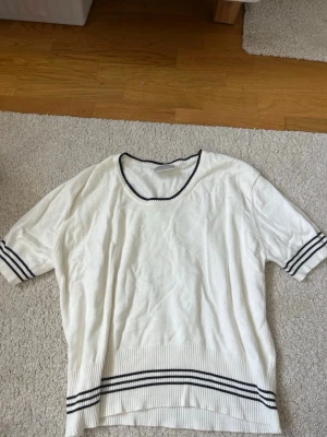 Vit topp med svarta ränder - Vit topp med svarta ränder vid halsringning, ärmslut och nederkant. Modellen har korta ärmar och en rund hals. Perfekt för en stilren och sportig look.
