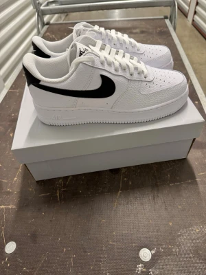 Nike Air Force 1 '07 Vita/Svarta Sneakers strl 43 - Klassiska Nike Air Force 1 sneakers i vitt läder med svart swoosh på sidorna. Skorna har perforerad tå för ventilation, vit sula och lågt skaft. Perfekta för dig som gillar en clean och tidlös look.
