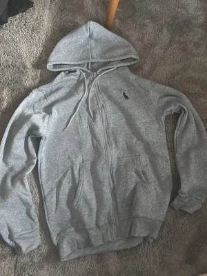 Grå zip hoodie - Snygg grå zip hoodie med klassisk huva och dragsko. Tröjan har dragkedja framtill, två fickor och den ikoniska mörkblå polospelaren broderad på bröstet. Tillverkad i mjukt bomullsmaterial som är skönt mot huden.