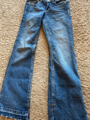 Blå bootcut jeans med slitna detaljer - Ett par blå bootcut jeans med slitna detaljer och råa kanter nertill. Jeansen har klassisk femficksdesign och ljusare partier framtill för en cool, avslappnad look. Perfekta för dig som gillar en retrostil och vill sticka ut.