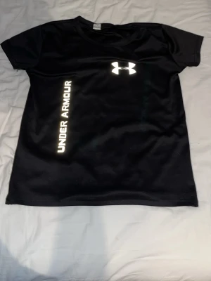 Svart t-shirt från Under Armour - Svart sportig t-shirt från Under Armour med vitt logotryck på bröstet och texten UNDER ARMOUR längs sidan. Tillverkad i ett lätt och snabbtorkande material som passar perfekt för träning och aktivitet. Klassisk rund hals och korta ärmar.