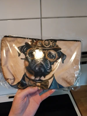 Necessär med steampunk-mopsmotiv - Necessär med beige bakgrund och tryck av en mops i steampunk-stil med hatt, glasögon och fluga. Väskan har dragkedja upptill och är tillverkad i tygmaterial. Perfekt för småprylar eller smink.