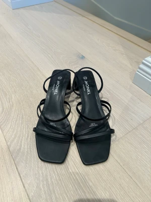 Svarta sandaler med klack från Monki - Snygga svarta sandaler från Monki med smala remmar och fyrkantig tå. Skorna har en medelhög blockklack och är tillverkade i veganskt material. Använda fåtal gånger.