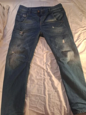 Blå slitna jeans med raka ben - Säljer ett par blå diesel jeans med snygga slitningar och fades med raka ben. Jeansen har klassisk femficksdesign, dragkedja och knapp i midjan. Materialet är denim i bomull och de har en avslappnad passform. Perfekta för en cool och avslappnad stil.