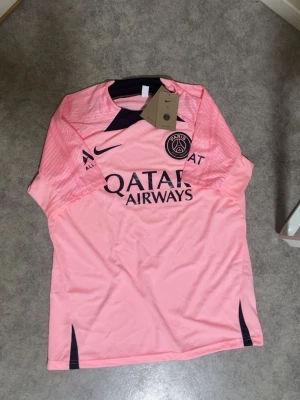 PSG rosa matchtröja från Nike - Säljer en snygg rosa Paris Saint-Germain matchtröja från Nike med svarta detaljer och klubbmärke på bröstet. Tröjan har Qatar Airways-tryck på framsidan och är tillverkad i ett lätt och ventilerande material, perfekt för fotboll eller träning.