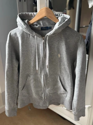 Grå hoodie från Polo Ralph Lauren - Grå zip-hoodie🧼 från Polo Ralph Lauren med vit broderad logga på bröstet. Klassisk modell med huva, dragsko och två fickor framtill. Mjuk bomullsblandning som känns skön mot huden. Perfekt för en avslappnad och clean look 🧼. Är öppen för att ändra priset 👀