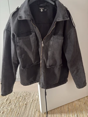 Svart jeansjacka från Boohoo - Cool svart jeansjacka från Boohoo med dragkedja, stora bröstfickor och axelremmar.  Grå/svart jeansfärg 