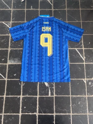 Sverige bortaställ VM 2026 | ISAK #9 - Säljer en blå Sverige fotbollströja från Adidas med gult tryck, nummer 9 och namnet Isak på ryggen. Tröjan har gula detaljer, klassiskt mönster och tre ränder på axlarna. Perfekt för dig som vill stötta landslaget med stil.