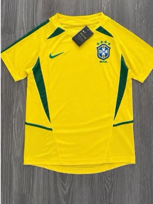 Brasilien Nike fotbollströja gul - Gul Brasilien fotbollströja från Nike med gröna detaljer och landslagets emblem på bröstet. Tröjan har korta ärmar, rund hals och är tillverkad i ett lätt, ventilerande material som passar perfekt till träning eller match.