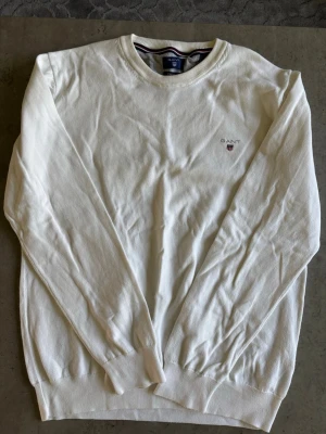 Vit tröja från GANT med broderad logo - Klassisk vit långärmad tröja från GANT med rund halsringning och diskret broderad logotyp på bröstet. Tröjan har ribbade muddar vid ärmslut och nederkant. Perfekt för en clean och stilren look.