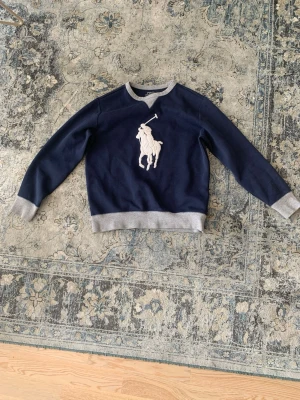 Blå sweatshirt från Polo Ralph Lauren - Mörkblå sweatshirt från Polo Ralph Lauren med grå muddar och krage. Stor vit broderad logga med polospelare på bröstet. Klassisk och sportig stil, perfekt till jeans eller joggers. Mjuk bomullsblandning för extra komfort. Det står ingen exakt storlek men den borde passa bra om du är runt 160-165 cm