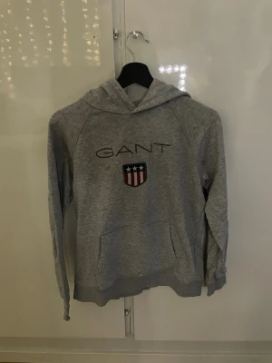 Grå hoodie från GANT med logga - Finns några fläckar. Grå hoodie från GANT med klassisk logga på bröstet. Hoodien har luva, känguruficka och ribbade muddar. 