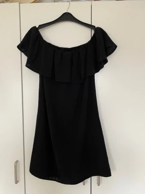 Svart offshoulder kortklänning - Snygg svart kortklänning med offshoulder-modell och bred volang upptill. Klänningen har en luftig passform och är perfekt för varma dagar. 