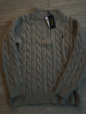 Beige kabelstickad tröja Polo Ralph Lauren - Kabelstickad beige tröja från Polo Ralph Lauren med rund halsringning och ribbade muddar. Tröjan har det klassiska broderade logotypen på bröstet och är tillverkad i ett mjukt, värmande material. Perfekt för lager-på-lager-stil.