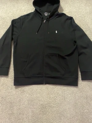 Svart kofta från Polo Ralph Lauren - Svart kofta med dragkedja från Polo Ralph Lauren. Den är i storlek Xl! Klassisk design med vit broderad logga på bröstet, huva med dragsko och två fickor framtill. Perfekt för en chill och stilren look.