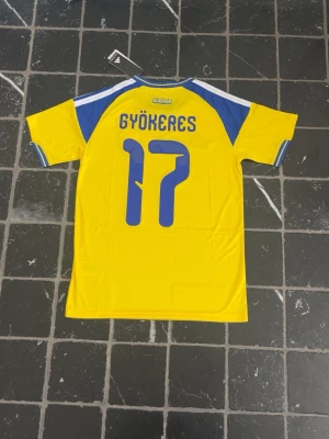 Svensk landslagströja Gyökeres 17 - Gul och blå fotbollströja från Adidas med Sveriges landslagsemblem och nummer 17 Gyökeres på ryggen. Tröjan har korta ärmar, blå detaljer på axlar och ärmslut samt tryckt logga och nummer. Perfekt för dig som vill stötta Sverige på match!