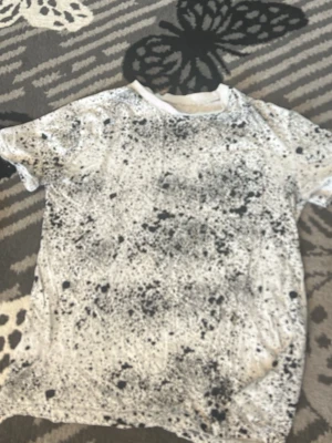Vit t-shirt med svart stänkmönster - Cool vit t-shirt med unikt svart stänkmönster över hela plagget. Klassisk passform med korta ärmar och rund halsringning. Perfekt för dig som vill sticka ut med en kreativ och modern stil.