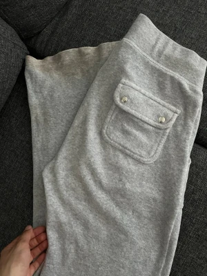 Grå mjukisbyxor med snörning - Snygga grå mjukisbyxor från Juicy Couture med snörning i midjan och stora fickor fram. Bakficka med lock och silverfärgade detaljer. Perfekta för en chill och avslappnad stil.