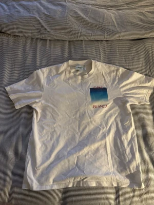 Vit Casablanca t-shirt med blått tryck - Vit t-shirt från Casablanca med ett stort blått och turkost tryck på ryggen och mindre tryck på bröstet. Röd text 'CASA BLANCA' både fram och bak. Klassisk passform och rund hals. Perfekt för en chill och stilren look.