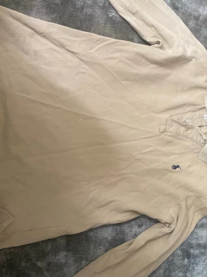 Beige långärmad polotopp från Polo Ralph Lauren - Klassisk beige långärmad polotopp från Polo Ralph Lauren med broderad logga på bröstet. Topp med krage och knappar framtill, tillverkad i bomull. Perfekt för en stilren och avslappnad look. Finns typ 2-3 fläckar av klorin på ryggen men dem är inte så stora o man märker knappt 
