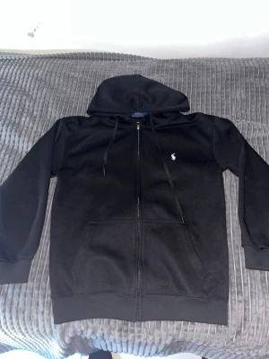 Svart hoodie från Ralph Lauren - Svart hoodie med dragkedja från Ralph Lauren. Klassisk design med huva, dragsnören och två fickor framtill. Liten vit broderad logga på bröstet. Perfekt för en chill och stilren look.