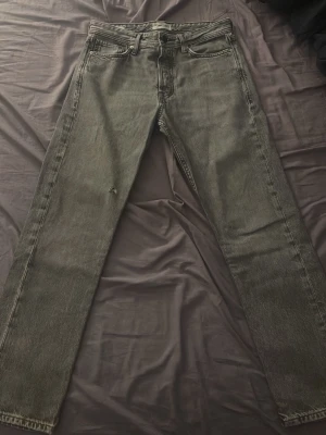 Grå raka jeans från Jack & Jones - Snygga grå jeans från Jack & Jones med rak passform och klassisk femficksdesign. Jeansen har en diskret slitning på ena benet och en läderpatch med logga bak i midjan. Tillverkade i denim med en cool vintagekänsla.