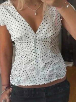 Vit prickig topp med v-ringning - Snygg vit topp med svarta prickar och djup v-ringning. Korta ärmar med lätt volang och knappar framtill. Figurnära passform som framhäver midjan. Perfekt till jeans eller kjol för en trendig look.