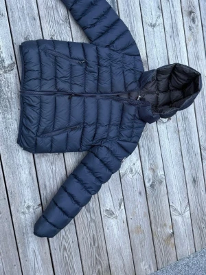 Colmar dunjacka - Snygg mörkblå dunjacka med quiltad design och huva. Jackan har långa ärmar, dragkedja framtill och två snedställda fickor med dragkedja. Perfekt för kyliga dagar och enkel att matcha med olika outfits. Är även en lagning på baksidan. Storlek 14 y 166cm pris kan gå ner vid snabb affär…