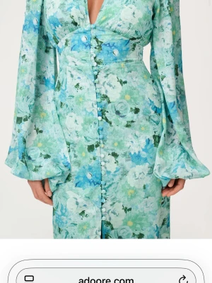 Blommig långklänning från Adoore - Långklänning från Adoore i ett blommigt mönster i gröna, blå och vita toner. Klänningen har långa, vida ärmar, markerad midja och knappar framtill och djup v-ringning .
