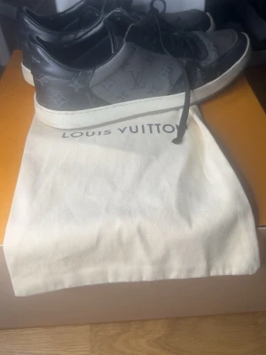 Louis Vuitton svarta sneakers - Snygga svarta sneakers från Louis Vuitton med klassiskt monogrammönster i grått och svart. Skorna har lågt skaft, svarta skosnören och vit sula. Tillverkade i läder och canvas för en lyxig känsla. Perfekta för dig som vill sticka ut med exklusiva detaljer.