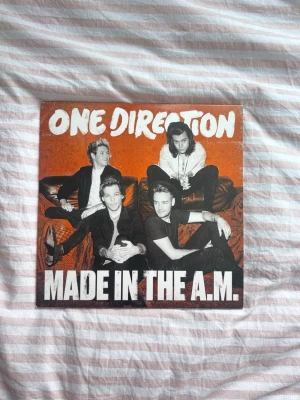 One Direction Made in the A.M. vinyl - Vinylskiva med albumet 'Made in the A.M.' av One Direction. Omslaget är orange med vit text och har ett foto på bandmedlemmarna på framsidan. Baksidan visar låtlistan och ett svartvitt foto av bandet. Perfekt för samlare eller fans av popmusik.