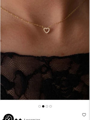Guldhalsband med hjärthänge - Ett elegant halsband i guld med ett hjärformat hänge dekorerat med små gnistrande pärlor. Kedjan är tunn och stilren, perfekt för att ge en söt touch till din outfit. Hjärtat är öppet i mitten och ger ett modernt och romantiskt intryck.
