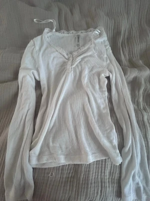 Vit ribbad långärmad topp H&M - Vit långärmad topp från H&M med ribbad struktur och spetsdetalj vid v-ringad hals. Smal passform och mjukt material som känns skönt mot huden. Perfekt att styla med jeans eller kjol för en enkel och snygg look.