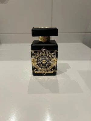 Initio Oud For Greatness Parfym - Lyxig parfymflaska från Initio med namnet Oud For Greatness. Flaskan är svart med detaljer i guld och har ett mystiskt ögonmotiv på framsidan. Kommer i en elegant svart ask med guldfärgat mönster. Perfekt för dig som gillar exklusiva dofter.
