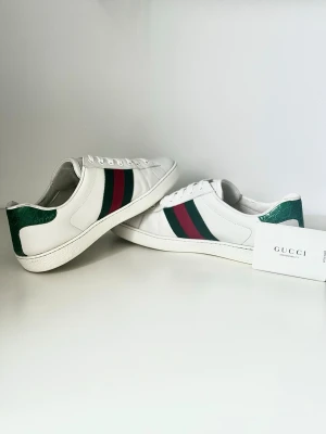 Gucci vita sneakers med gröna detaljer - Säljer ett par klassiska vita sneakers från Gucci med gröna och röda ränder på sidan samt grön hälflik i strukturmönstrat läder. Skorna har lågt skaft, vita snören och är tillverkade i slätt läder. Perfekta för dig som gillar exklusiv och tidlös stil. Äkthetsbevis medföljer.