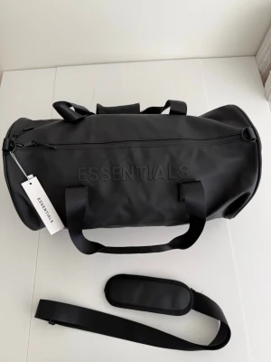 Svart duffelbag från Essentials - Snygg svart duffelbag från Essentials med stilren design och tydlig logga på sidan. Väskan har robusta handtag, avtagbar axelrem och dragkedja från YKK. Tillverkad i ett tåligt, vattenavvisande material som passar perfekt för resor eller gymmet.