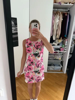 Blommig rosa kortklänning - Snygg kort klänning med blommigt mönster i rosa, svart och vitt. Ärmlös modell med rund halsringning och figurnära passform. Perfekt för varma dagar och somriga tillfällen.