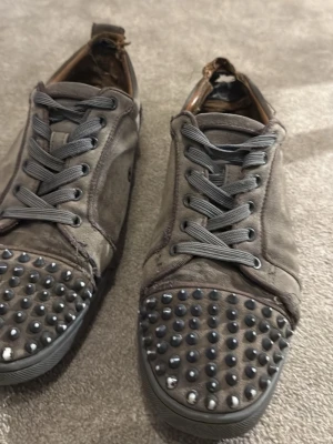 Grå sneakers med nitar på tån - Grå loubs som är slitna men går att fixa. Unika grå sneakers med coola nitar på tån och grå snörning. Skorna har en låg profil och är tillverkade i mockaliknande material med detaljer i läder. Perfekta för dig som vill sticka ut med din stil.