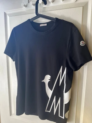 Svart Moncler t-shirt ( äkta ) - Svart t-shirt från Moncler med stort vitt tryck av logga och tupp på framsidan samt texten 'MONCLER' på baksidan. Klassisk rund halsringning och korta ärmar. Perfekt för en trendig och avslappnad stil. Ny pris:2900
