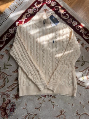 Polo Ralph Lauren Cable-Knit Quarter-Zip (Storlek S) - Kabelstickad cremevit tröja från Polo Ralph Lauren med halv dragkedja och hög krage. Tröjan har ribbade muddar och nederkant samt den klassiska broderade loggan i marinblått på bröstet. Perfekt för en stilren och avslappnad look.