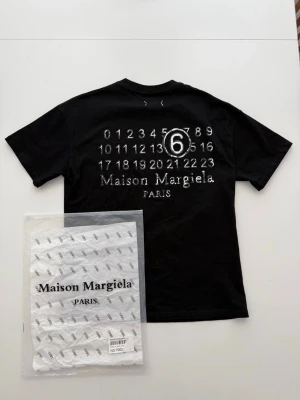 Maison Margiela svart t-shirt - Maison Margiela svart t-shirt med ikoniskt tryck av siffror och logga på ryggen. Klassisk passform och rund hals. Tillverkad i mjuk bomull för en skön känsla. Perfekt för dig som gillar streetwear och vill sticka ut med en unik design. Pris kan diskuteras💸 Strl:S✅ Hör av dig om du har några funderingar!👍 Aldrig använd!🤩